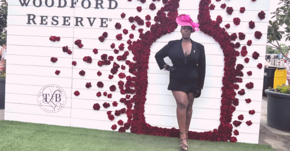 Stylish Black woman poses confidently at the Kentucky Derby in a chic black blazer dress and bold pink fascinator. Surrounded by elegant red rose decor at the Woodford Reserve lounge, embodying race day fashion and southern charm with poise.