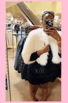 The Luxury Bandit wearing a chic white faux fur stole, taking a mirror selfie in a Dillard's Clearance Center, surrounded by racks of clothing, with The Luxury Bandit logo. This image showcases successful luxe for less shopping and finding Dillard's outerwear clearance gems.