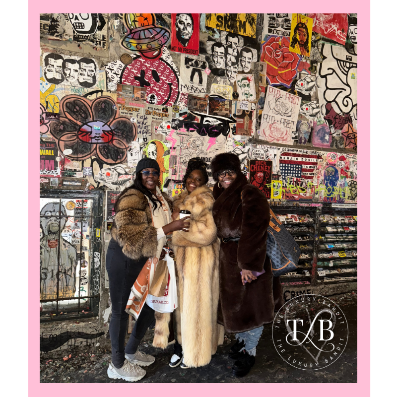 Three fashionable women wearing luxury fur coats and sunglasses pose together at Pike Place Market in Seattle, standing in front of colorful street art and graffiti-covered walls in the famous Pacific Northwest destination