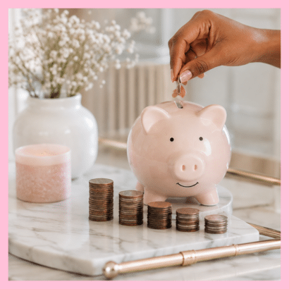 A hand adds a coin to a blush pink piggy bank beside growing stacks of coins on a marble surface, creating a visual reminder of how consistent contributions can build wealth over time. This feminine savings scene represents retirement planning, 401(k) contributions, and compound growth while showing how small financial decisions today can support a stronger financial future.